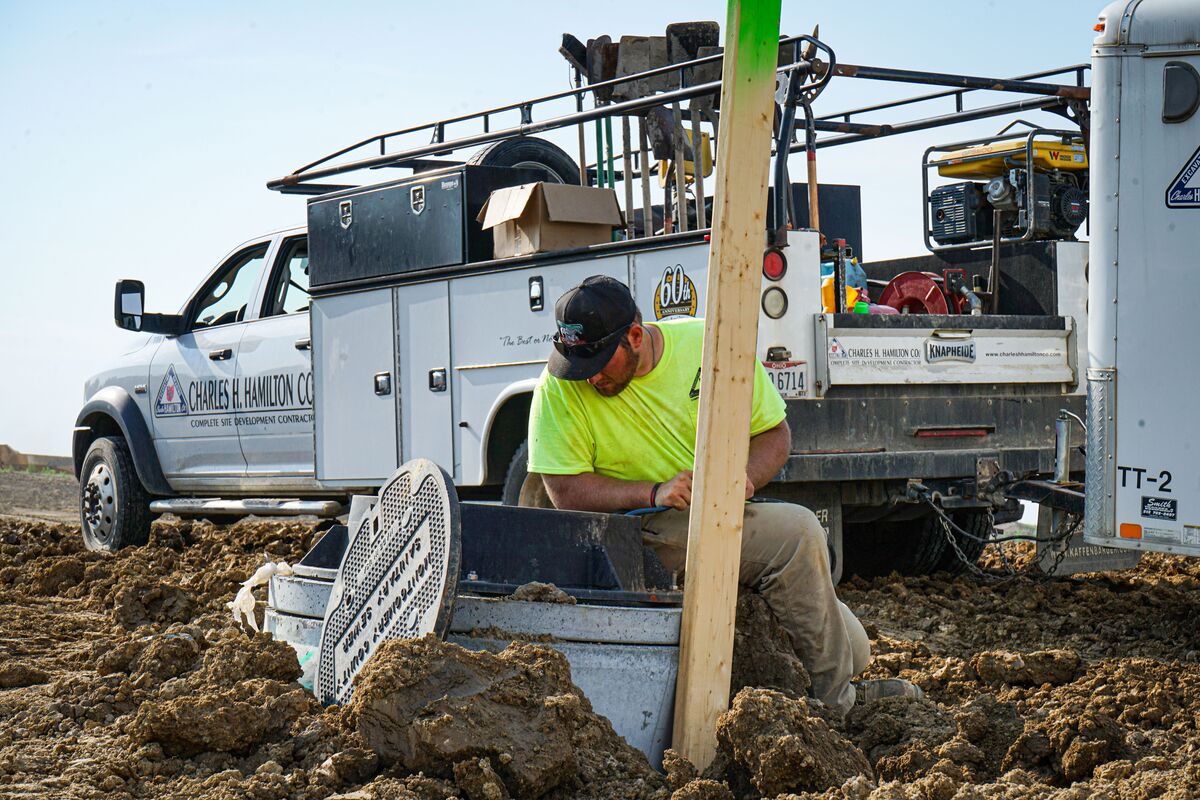 Construction worker kneeling in muddy terrain while performing utility or site development work beside a service truck loaded with tools and equipment.