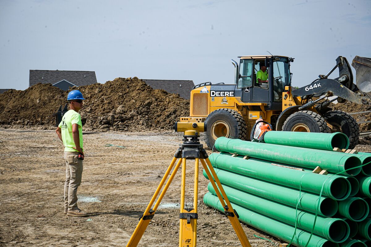 construction worker in blue hard hat standing in front of pile of pipes and wheel loader
