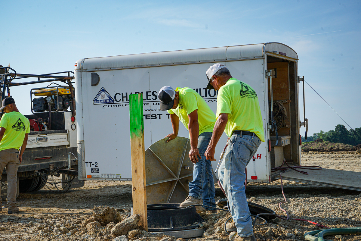 Construction crew installing a utility access structure beside a service trailer at an active job site.