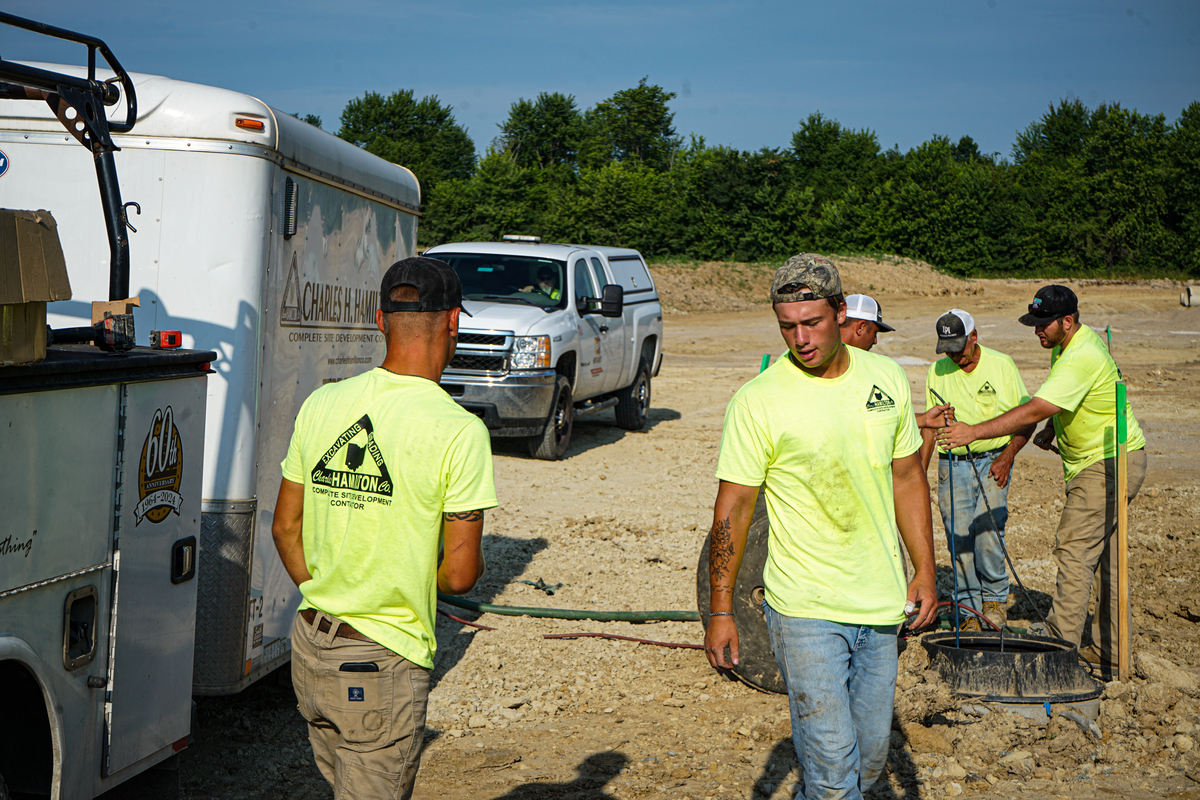 Construction crew in high-visibility shirts working on a utility installation at an active job site, with service trucks, equipment, and team members coordinating in the background.