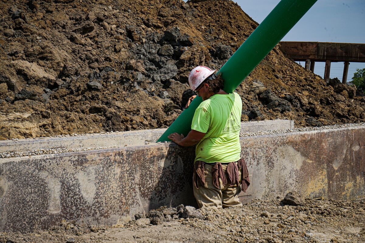 Construction worker carrying a large green pipe while standing in a trench at an active utility installation site.